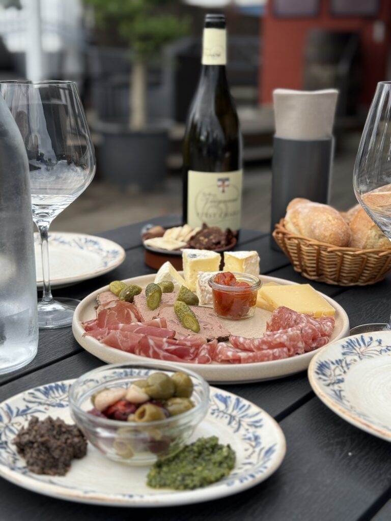 Tapasplatte med skinke, ost og tilbehør serveret med hvidvin og friskbagt brød.